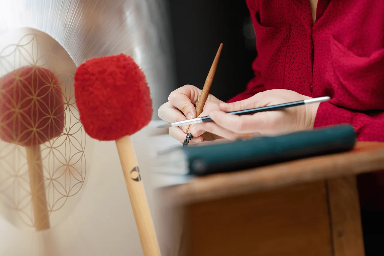 Zwei Gong-Schlägel vor einer Blume des Lebens, daneben eine Hand bei einer Kalligrafie